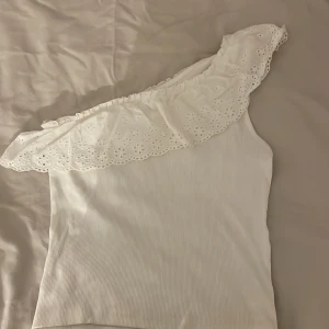 Vit oneshoulder topp från Kappahl - Vi säljer en vit oneshoulder topp från Kappahl med en vacker spetsvolang längs axeln. Toppen är ribbad och perfekt för sommarens varma dagar. Passar både till jeans och kjol för en stilren look. Den är använd ca 3-4 gånger och jag säljer den för att den är för liten!😽💗