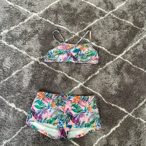 Färgglad blommig bikini - Säljer en superfin bikini med tropiskt blommönster i rosa, grönt och lila. Toppen har smala axelband och byxorna är i shortsmodell. Perfekt för sommarens stranddagar! Testad.