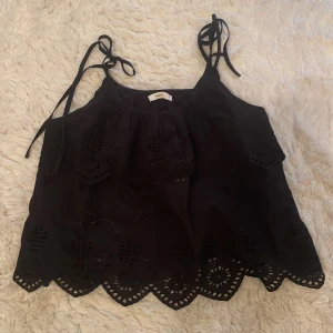 Svart ärmlös topp från Pigalle - Säljer en svart ärmlös topp från Pigalle med broderade detaljer och knytband vid axlarna. Toppen har en vågig kant och är perfekt för varma dagar.