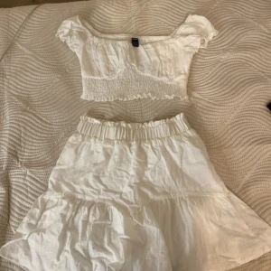 Två delad , Vit croppad topp och en vit kjol - Säljer en söt vit croppad topp från med korta ärmar och smockdetaljer. Toppen har en elastisk passform och volangkant. Kjolen har också en elastisk passform och volabgkant. Perfekt för varma sommardagar! Eller för skolavslutninge!