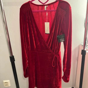 Röd omlottklänning från Nelly - Snygg röd omlottklänning i sammet från Nelly. Klänningen har långa ärmar och en v-ringad design med knytband i midjan. Perfekt för att sticka ut med sin djupa färg och mjuka material. Aldrig använd och prislappen sitter kvar!