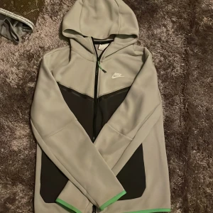 Grå hoodie från Nike - Säljer en grå hoodie från Nike med dragkedja och huva. Den har svarta detaljer och en vit Nike-logga 