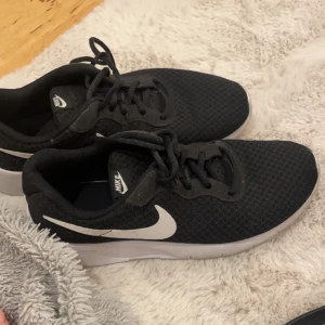 Svarta Nike sneakers - Säljer ett par svarta Nike sneakers med vit sula och klassisk Nike-logga på sidan. Skorna har snörning och är perfekta för en sportig look. De är bekväma och passar till många olika tillfällen.
