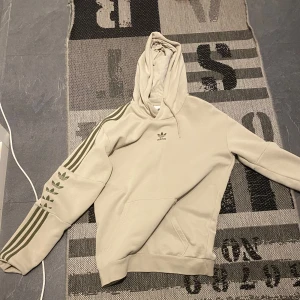 Beige hoodie från Adidas - Säljer en snygg beige hoodie från Adidas med klassiska tre ränder i grönt längs ärmarna. Hoodien har en känguruficka och en justerbar huva. Perfekt för en avslappnad stil.