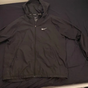 Svart vindjacka från Nike - Säljer en svart vindjacka från Nike med huva och dragkedja. Jackan har långa ärmar och två fickor framtill. Perfekt för löpning och andra utomhusaktiviteter. Hör av er vid minsta lilla fundering 🙌😉