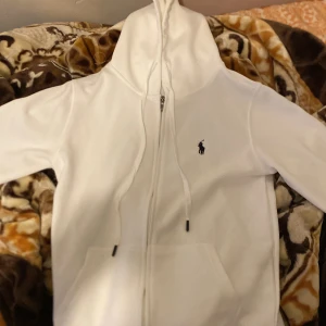 Vit hoodie från Polo Ralph Lauren - Säljer en stilren vit hoodie från Polo Ralph Lauren med dragkedja och det klassiska broderade logotypen på bröstet. Perfekt för en avslappnad look. Den har en justerbar huva och praktiska fickor framtill. Skick 10/10 pris kan förhandlas vid snabb äffär