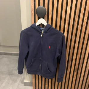 Mörkblå huvtröja från Ralph Lauren - Säljer en snygg mörkblå huvtröja från Ralph Lauren med dragkedja och en röd broderad logga på bröstet. Tröjan har en praktisk huva. Perfekt för en avslappnad stil. Lägsta pris 350kr.🙌