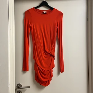Orange klänning från H&M - Snygg orange klänning från H&M med långärmad design och draperad detalj framtill. Perfekt för att sticka ut med sin livfulla färg och unika stil. Klänningen har en åtsittande passform som framhäver figuren.