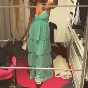 Pastell Grön långklänning med volanger - Säljer en vacker pastell grön långklänning med volanger. Klänningen har smala axelband och en luftig passform, perfekt för sommarens festligheter. Den har flera lager av volanger som ger en fin rörelse. Passar väldigt bra som balklänning. Små  hål längst ner men syns inte då volangen faller så fint