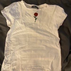 Vit t-shirt med rosdetalj - Säljer en vit t-shirt med en broderad röd ros på bröstet. T-shirten har korta ärmar och en klassisk rund halsringning. Perfekt för en stilren och enkel look.