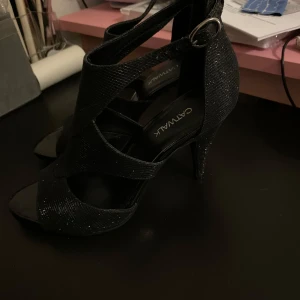 Svarta glittriga klackskor från Catwalk - Snygga svarta klackskor från Catwalk med glittrig yta och öppen tå. Skorna har en elegant design med remmar och en liten spänne på sidan. Perfekta för att ge en extra touch till din outfit.