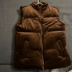 Brun quiltad väst - Säljer en snygg brun quiltad väst med dragkedja. Perfekt för kyligare dagar och ger en stilren look. Västen har en hög krage och två praktiska fickor framtill.