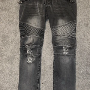 Grå jeans med dragkedjor från Balmain - Snygga grå jeans från Balmain med slitna detaljer och dragkedjor vid fickorna. Byxorna har en tight passform och ribbade partier vid knäna för en cool look. Perfekta för dig som vill ha en edgy stil.