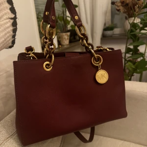Michael Kors  - Säljer min Michael Kors Cynthia Saffiano Leather Satchel i färgen burgundy. Väskan är i mycket gott skick och har välvårdad handtag. Det finns en liten misfärgning på kedjan, men den syns knappt och påverkar inte väskans utseende i övrigt. Inga repor