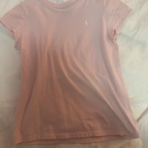Rosa t-shirt från Ralph Lauren - Säljer en snygg rosa t-shirt från Ralph Lauren med det klassiska broderade logotypen på bröstet. T-shirten är kortärmad och tillverkad i mjuk bomull. Perfekt för en avslappnad stil. Tröjan är i storlek L men det är barnstorlek så tröjan passar mig som har storlek XS och S normalt.