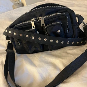 Svart axelväska med nitar nunoo  - Snygg svart axelväska med flera dragkedjor och coola nitar på axelremmen. Perfekt för att ge din outfit en edgy touch. Väskan har flera fack för enkel organisering och en justerbar rem för bekväm passform.