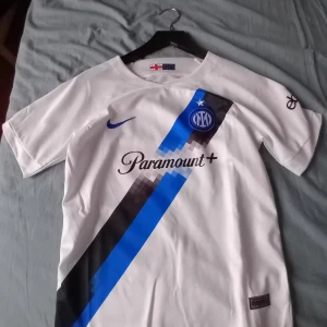 Vit fotbollströja från Nike med Inter-logga - Säljer en snygg vit fotbollströja från Nike med Inter-logga. Tröjan har korta ärmar och ett coolt diagonalt mönster i blått och svart. Den har också sponsorer som Paramount+ och eBay tryckta på framsidan och ärmen. Perfekt för alla Inter-fans!