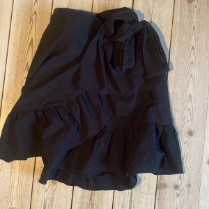 Svart kjol från Vero Moda - Säljer en superfin svart kjol från Vero Moda. Den har en omlott-design med volangdetaljer och knytning i midjan. Perfekt för både vardag och fest! 🖤
