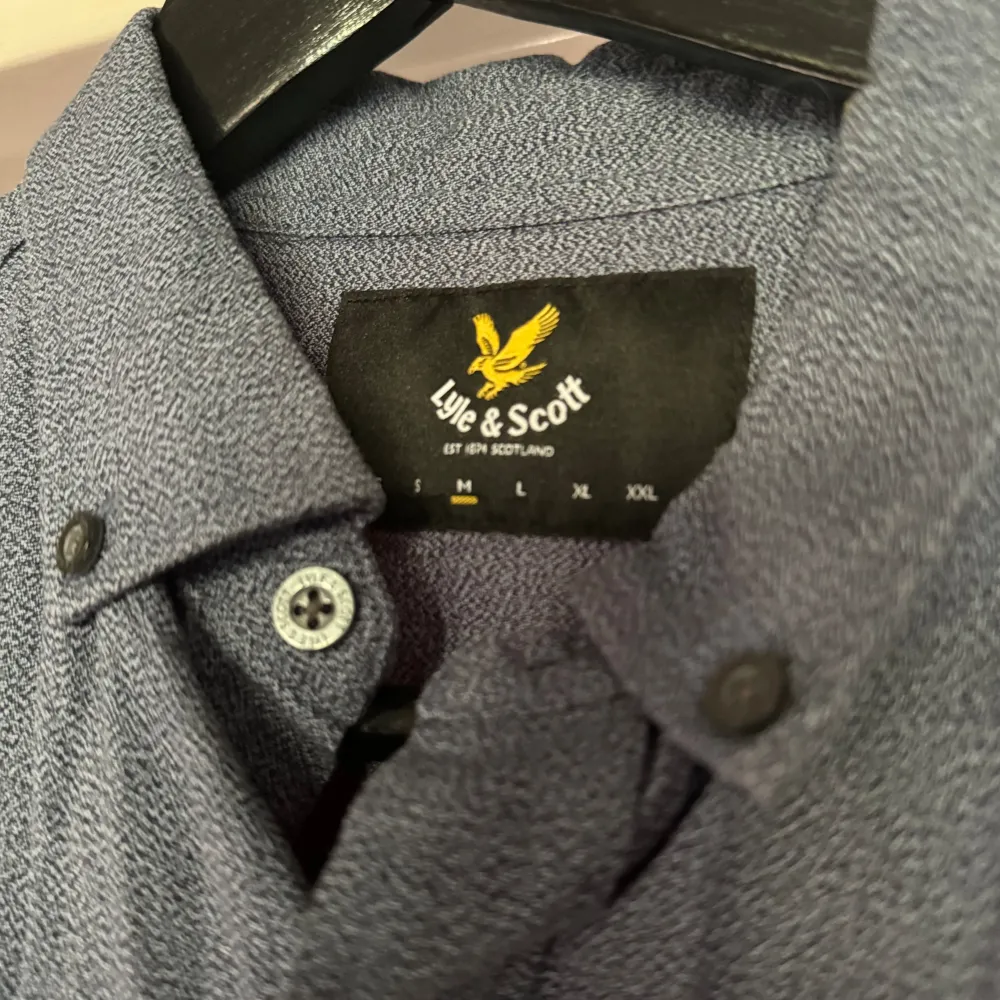 Säljer en snygg blå skjorta från Lyle & Scott. Den har en klassisk krage och långa ärmar, perfekt för både vardag och fest. Skjortan är dekorerad med det ikoniska gula logotypen på bröstet och har knappar hela vägen ner. Materialet känns bekvämt och stilrent.. Skjortor.