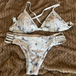 Vit och blå mönstrad bikini - Säljer en superfin vit och blå mönstrad bikini i storlek M. Den har ett coolt marmorerat mönster och smala axelband. Bikinitrosorna har snygga sidodetaljer med flera band. Perfekt för sommarens strandhäng eller poolpartyn!