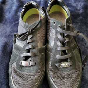 Svarta sneakers från Hugo Boss - Säljer ett par stilrena svarta sneakers från Hugo Boss. Skorna har en snygg kombination av mocka och textilmaterial med klassisk snörning. De är i storlek herr 43 och har en diskret logga på sidan. Perfekta för både vardag och fest!