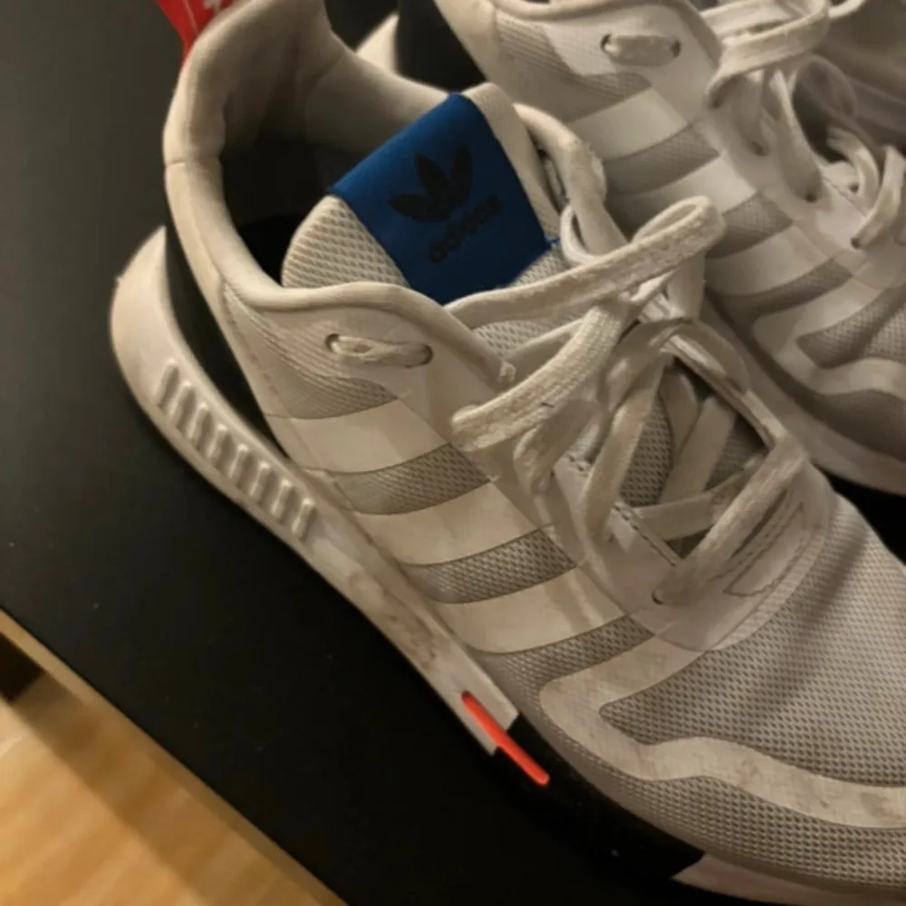 Säljer ett par snygga vita Adidas sneakers med klassiska tre ränder på sidan. Skorna har en blå detalj på plösen och en liten orange accent på sulan. De är i bra skick och perfekta för både vardag och träning.. Skor.