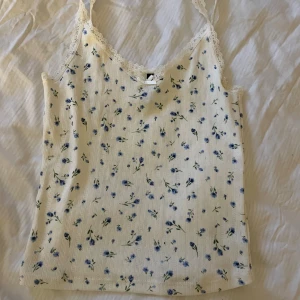 Blommig linne från H&M Divided - Säljer ett supersött linne från H&M Divided i storlek XS. Det är vitt med ett blommigt mönster i blått och grönt. Linnet har spetsdetaljer vid halsringningen och en liten rosett framtill. Perfekt för sommardagar!
