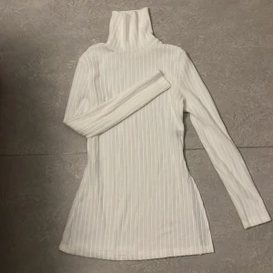 Vit ribbad polotröja - Säljer en stilren vit ribbad polotröja med långa ärmar. Perfekt för kyligare dagar och enkel att matcha med olika outfits. Tröjan har en klassisk polokrage som ger extra värme och stil.