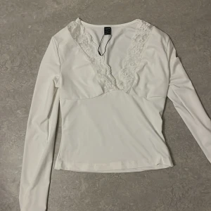 Vit långärmad topp med spetsdetaljer - Säljer en stilren vit långärmad topp från H&M med vackra spetsdetaljer längs halsringningen. Perfekt för en elegant look. Toppen har en figurnära passform och är tillverkad i ett mjukt material.