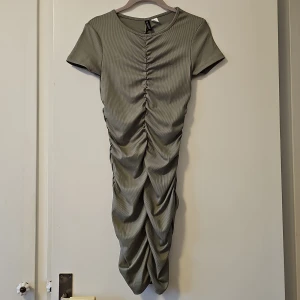 Grön klänning från H&M Divided - Snygg grön klänning från H&M Divided med rynkade detaljer framtill och baktill. Klänningen har korta ärmar och är tillverkad i en mjuk polyesterblandning med elastan för extra stretch. Perfekt för en stilren look!