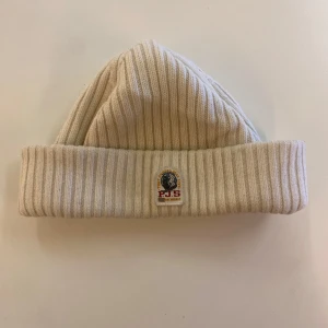Beige ribbad mössa från Parajumpers - Snygg beige mössa från Parajumpers med ribbad design och uppvikt kant. Perfekt för kyliga dagar med stilren look. Märkeslogga framtill ger en exklusiv touch. Lite missfärgad men ändå fint skick