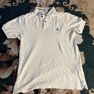 Vit pikétröja från Stone Island - Säljer en stilren vit pikétröja från Stone Island. Tröjan har en klassisk krage med knappar och en broderad logotyp på bröstet. Perfekt för en avslappnad och snygg look.