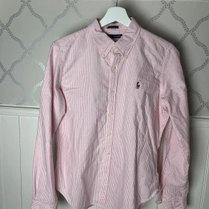 Rosa och vit randig skjorta från Ralph Lauren - Snygg rosa och vit randig skjorta från Ralph Lauren med klassisk krage och knappar framtill. Skjortan har långa ärmar och en broderad logotyp på bröstet. Perfekt för en stilren look! (Liten reva på tredje bilden)