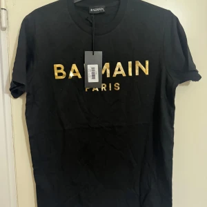 Svart t-shirt från Balmain - Snygg svart t-shirt från Balmain med guldig text på bröstet. Perfekt för en stilren look. Kortärmad och i mjukt material.
