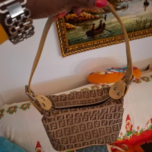 Beige och brun axelväska med monogram - Snygg axelväska i beige och brun med monogrammönster. Väskan har en justerbar axelrem och stängs med dragkedja. Perfekt för att ge en stilfull touch till din outfit.