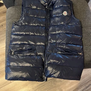 Mörkblå dunväst från Moncler - Säljer en snygg mörkblå dunväst från Moncler med dragkedja och två fickor framtill. Västen har en glansig finish och Moncler-logga på bröstet. Perfekt för kyligare dagar!