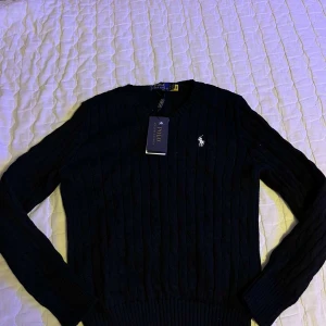 Svart stickad tröja från Polo Ralph Lauren - Säljer en stilren svart stickad tröja från Polo Ralph Lauren med det klassiska broderade logotypen på bröstet. Tröjan har en ribbad struktur och långa ärmar, perfekt för kyligare dagar. Ett måste i garderoben för den modeintresserade! 🖤