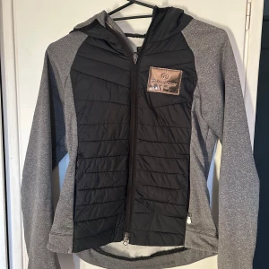 Grå och svart jacka med huva - Snygg grå och svart jacka från JH COLLECTION med huva och dragkedja. Jackan har en quiltad framsida. Perfekt för kyligare dagar.