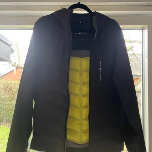 Svart jacka från Sail Racing - Snygg svart jacka från Sail Racing med huva och dragkedja. Den har en stilren design med en logotyp på bröstet och ryggen. Perfekt mot regn samt vind, Storlek XS. Nästintill oanvänd