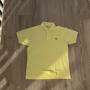 Gul pikétröja från Lacoste - Säljer en klassisk gul pikétröja från Lacoste med den ikoniska krokodilloggan på bröstet. Tröjan har korta ärmar och en traditionell krage med knappar. Perfekt för en stilren och avslappnad look.