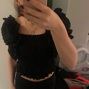 Svart volangtopp från Polio & Co - Säljer en svart volangtopp från Polio & Co. Toppen har en smockad design med volanger vid ärmarna och nederkanten. Perfekt för en trendig look. Jag har S och passar mig super!