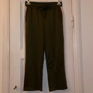 Byxor  - Snygga olivgröna byxor med vita ränder på sidan. Byxorna har en rak och avslappnad stil, perfekta för en casual look. Tillverkade i ett mjukt och skönt material.