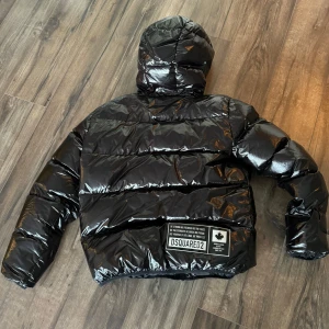 Svart glansig pufferjacka från Dsquared2 - Snygg svart glansig pufferjacka från Dsquared2 med huva och dragkedja framtill. Jackan har en cool logga på bröstet och en större patch på ryggen. Perfekt för att hålla värmen med stil.
