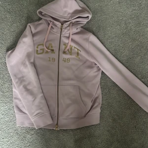 Ljusrosa hoodie från GANT - Säljer en snygg ljusrosa hoodie från GANT med dragkedja och justerbar huva. Tröjan har en stor GANT-logga och årtalet 1949 tryckt på framsidan. Perfekt för skolan eller om man bara vill stanna hemma. Den passar som både M och S 
