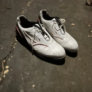 Vita fotbollsskor med snörning - Säljer ett par vita golfskor från mizuno med snörning och svarta sulor. Skorna har röda detaljer och en modern design.
