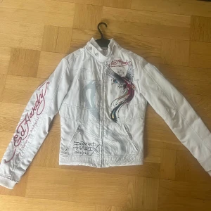 Vit jacka från Ed Hardy - Säljer en cool vit jacka från Ed Hardy med färgglada broderier av en fågel och en tiger. Jackan har långa ärmar och dragkedja framtill. Den är dekorerad med Ed Hardy-loggor i rött på ärmarna och ryggen. Skriv vid frågor eller diskutering av pris! Köpte på loppis så kan ej försäkra att den är äkta till 100 procent men allt tyder på det. Känns exklusiv i materialet och är extremt sparsamt använd.