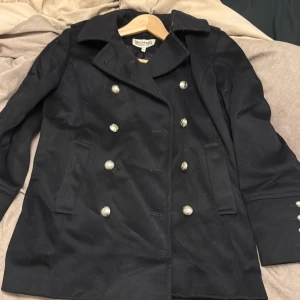 Svart kappa från Michael Kors - Snygg svart kappa från Michael Kors med dubbelknäppning och stiliga guldknappar. Den har en klassisk krage och är fodrad för extra komfort. Perfekt för kyligare dagar när du vill se extra elegant ut. Perfekt för tidig vår.