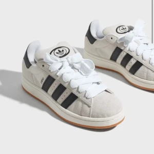 Adidas Campus 00s W, strl 40 - Snygga Adidas Campus sneakers i beige mocka med svarta detaljer och klassiska tre ränder. Skorna har en vit sula och snörning framtill. Perfekta för en stilren och avslappnad look. Använda 2 gånger så i nyskick. Nypris 1449kr. Storlek 40, tycker själv att adidas skor är något små i storleken. Betalning sker via Swish och skickas med PostNord. 