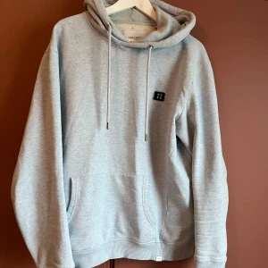 Ljusgrå/blå hoodie från Les Deux - Säljer en ljusgrå/blå hoodie från Les Deux. Den har en klassisk design med dragsko i huvan och en liten logotyp på bröstet.