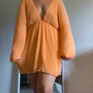 Orange klänning med v-ringning - Säljer en snygg orange klänning med v-ringning och långa ärmar. Klänningen har en luftig passform och knytning i ryggen för en extra detalj. Perfekt för sommarens varma dagar.